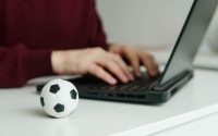 Young man reading soccer news or writing football article on laptop. Soccer ball on the table. Betting, gambling concept.